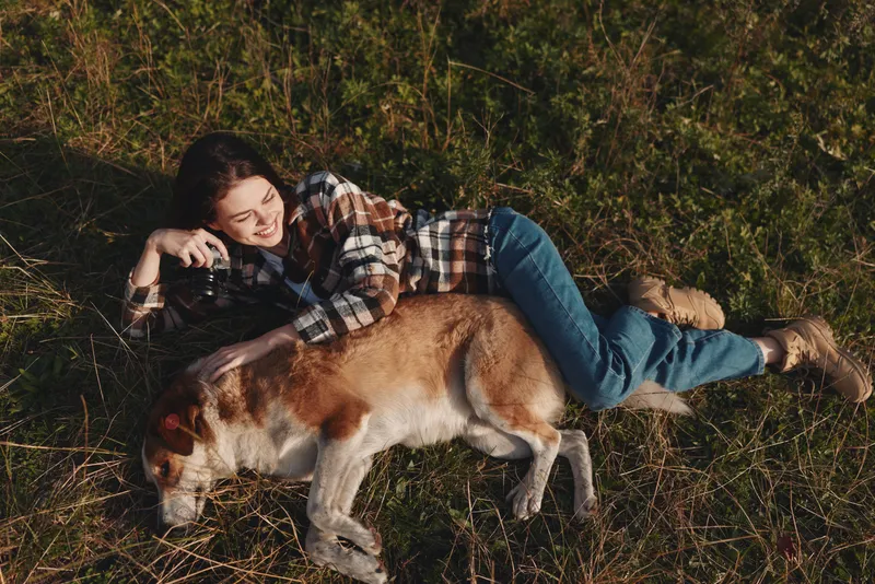 Woman lying on grass with a dog, camera in hand, capturing a candid outdoor moment during leisure time in a sunny field, relaxed atmosphere and strong companionship