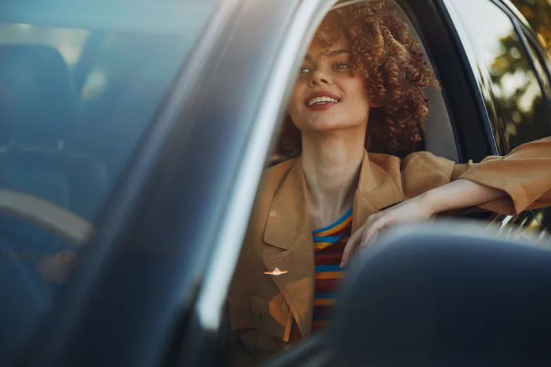 Woman in a car, smiling softly through the window, sunlit moment, relaxed pose, warm retro film color, casual blazer, striped top, intimate lifestyle portrait with natural light