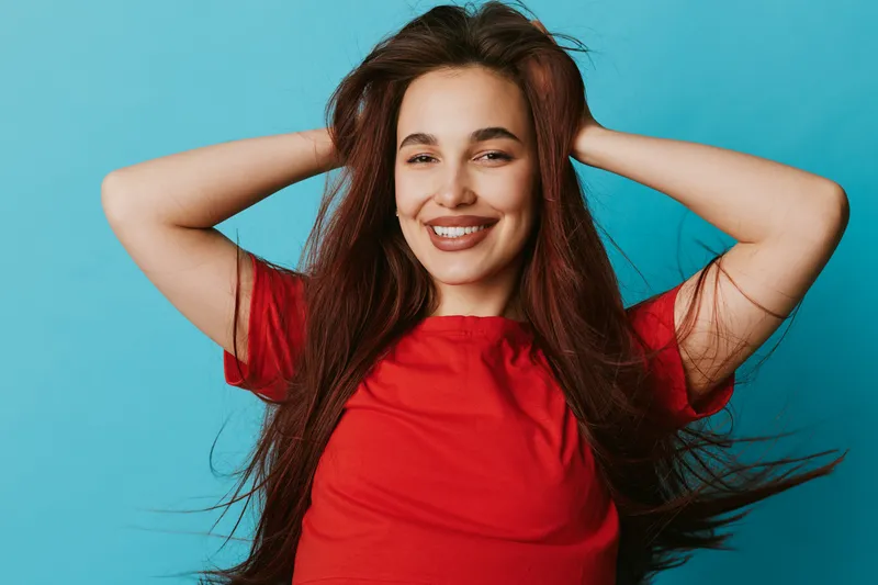 Young woman with long brown hair smiling and posing with hands behind head on blue background. Casual style, happy expression and natural beauty in studio portrait.