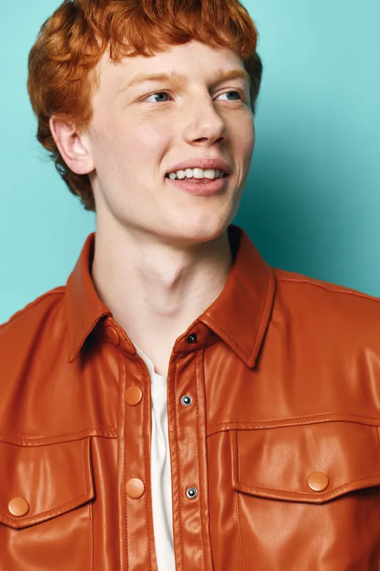 Young man with red hair wearing brown leather jacket and white shirt smiling and looking away on blue background. Casual style and happy expression in studio portrait photography.