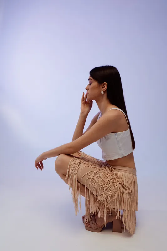 Young woman in a white top and fringed skirt poses in profile sitting on heels on a light background. Concept of beauty, fashion, and style.