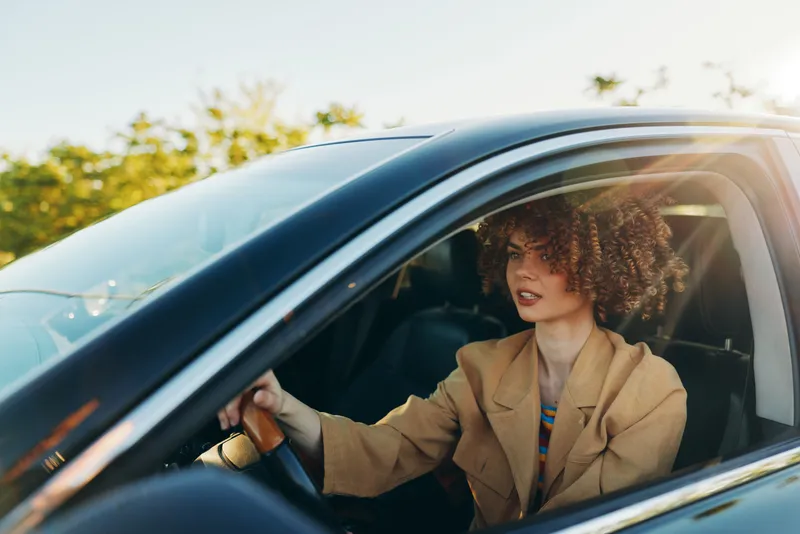 Bright scene shows a woman driving a car, casual beige coat, sunlit window, retro color palette, relaxed mood, natural tones, candid lifestyle moment, smiling vibe, urban road