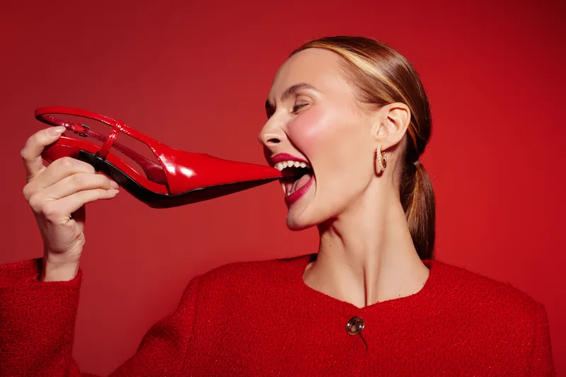 A smiling woman in red sweater holds a shiny red high heel shoe near her mouth against a red background. Concept for fashion, style, beauty, and bold color contrast.