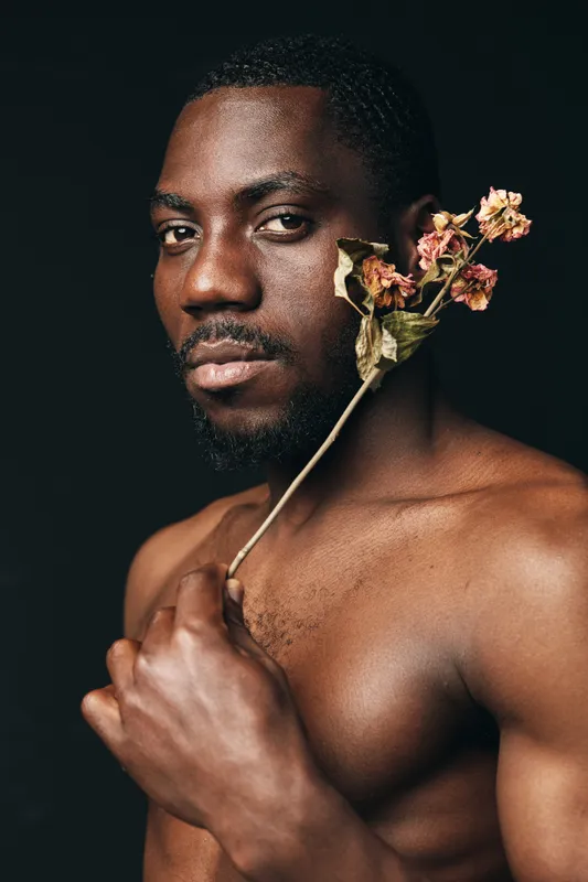 Black man with dark skin holding dried flowers near face in creative conceptual visual style portrait with serious expression on dark background.