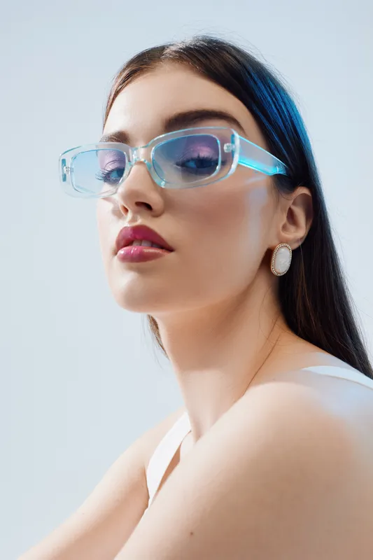 Young woman with long dark hair wearing transparent blue sunglasses and white top poses against light background. Concept of beauty, fashion, elegance, and modern style.