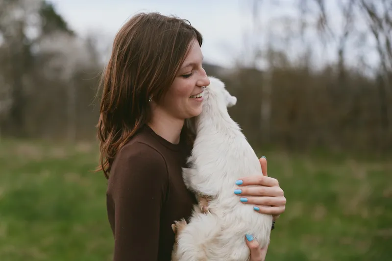 A woman in casual clothing hugs a white goat kid in an open field, smiling softly. A warm lifestyle moment highlighting animal bond, tenderness, and rural living.