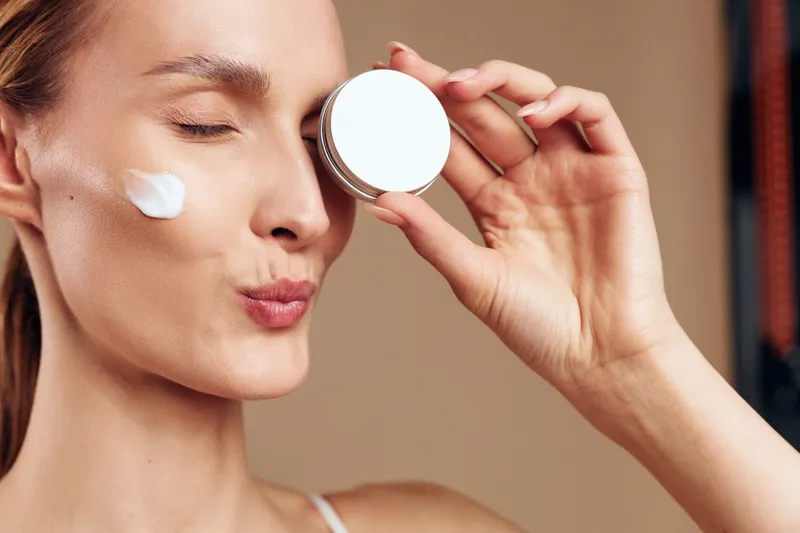 A young woman with clear skin applies face cream on her cheek while holding a small round container near her eye. The neutral background highlights skincare and beauty concepts.