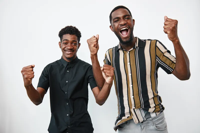 Two young black men with dark skin celebrating success with raised fists and smiling faces in casual clothing against white background in studio. Friendship and joy.