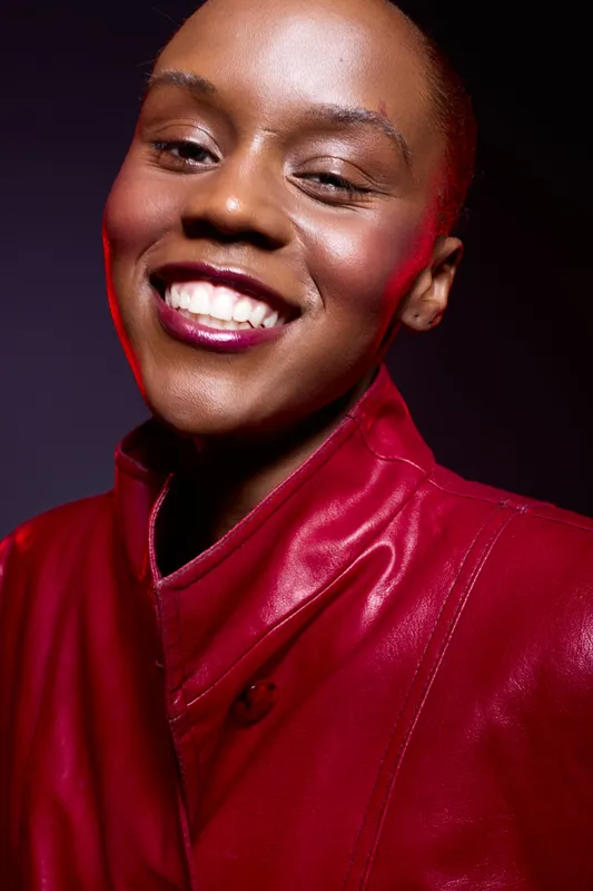 Close-up of a smiling young black woman with dark skin wearing a red leather jacket. Studio portrait with dramatic lighting on a dark background. Concept of beauty, fashion, and confidence.