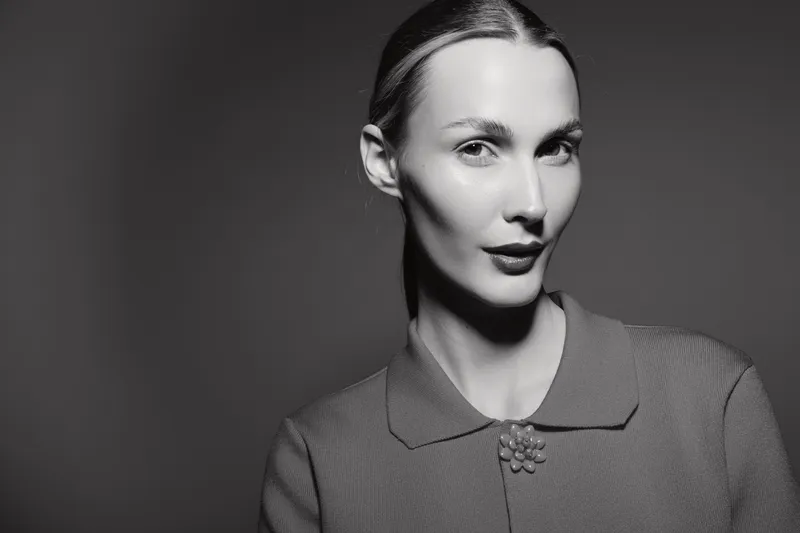 Portrait of a young white woman with fair skin and pulled-back hair wearing a textured coat with a brooch, posing against a dark neutral background in soft lighting.