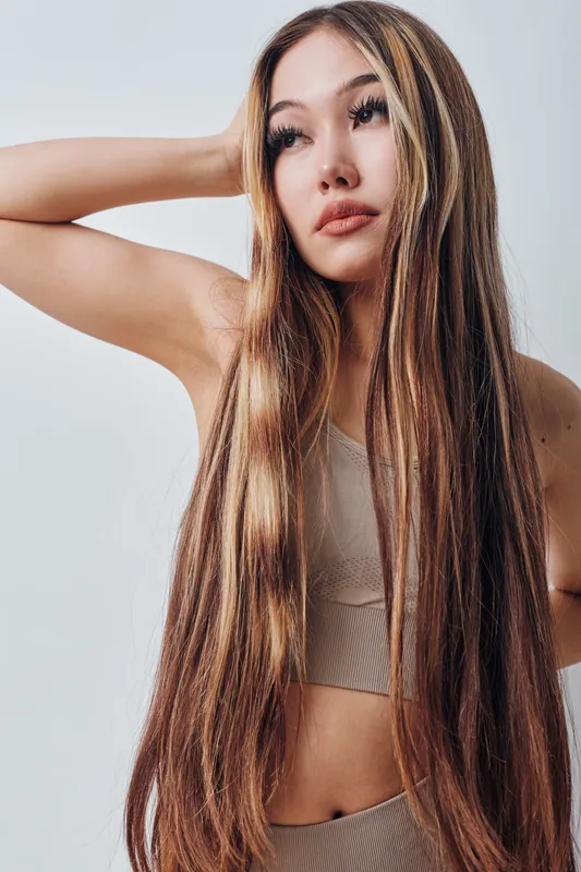 Woman with long straight hair posing with hand behind head looking away. Fashion model with healthy hairwearing beige outfit on light background in studio portrait.
