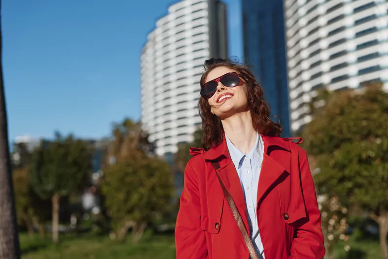 Young woman with curly hair wearing a red coat and sunglasses enjoys a sunny day outdoors with modern buildings in the background.