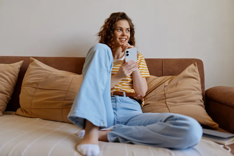 Smiling woman with curly hair sitting on a couch at home, wearing a striped shirt and jeans, holding a smartphone. Cozy living room vibe, relaxed mood, casual lifestyle.
