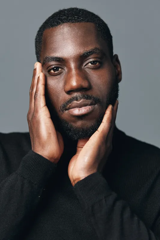 Portrait of African man with dark skin and beard in creative conceptual visual style wearing black sweater touching face with hands against gray background.