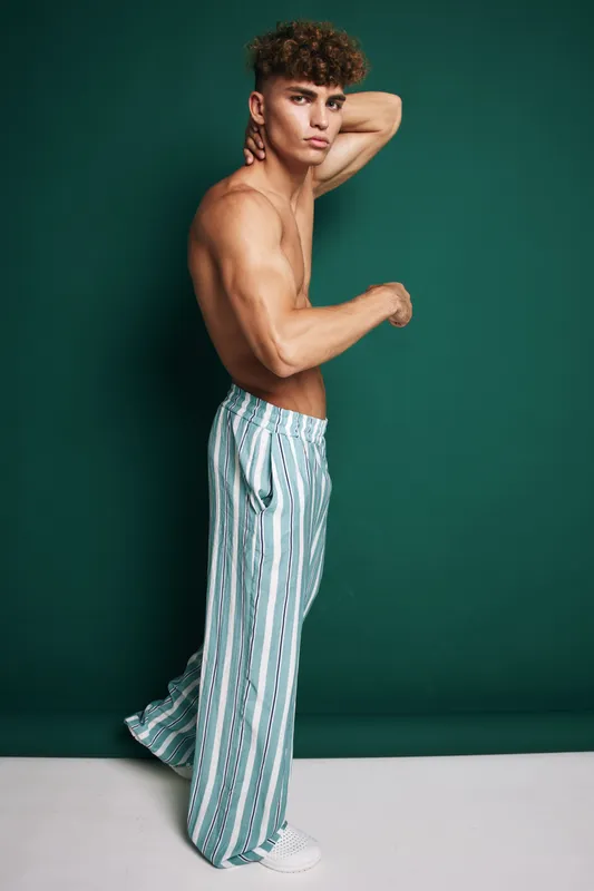 Young muscular man posing shirtless in striped wide pants and white sneakers against green background for fashion and fitness concept.