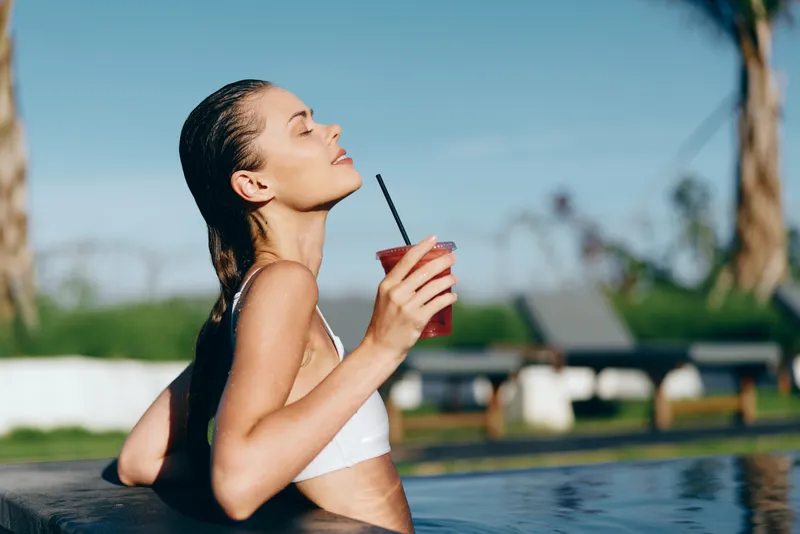Young woman with braided hair relaxes in an outdoor pool holding a refreshing drink with a straw while enjoying the sunny day with closed eyes and calm expression.