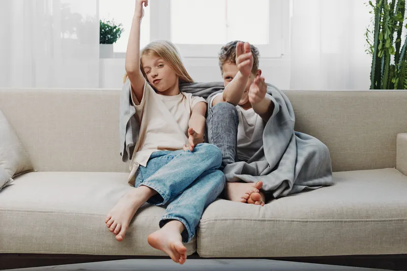 Two children sitting on a beige sofa wrapped in a gray blanket playing and relaxing in a bright living room with plants and large windows.