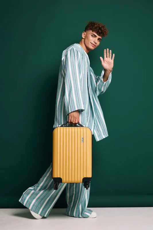 Young man in striped pajamas walking with yellow suitcase waving for hello on studio background. Casual style travel, luggage and friendly gesture with smile and curly hair portrait.