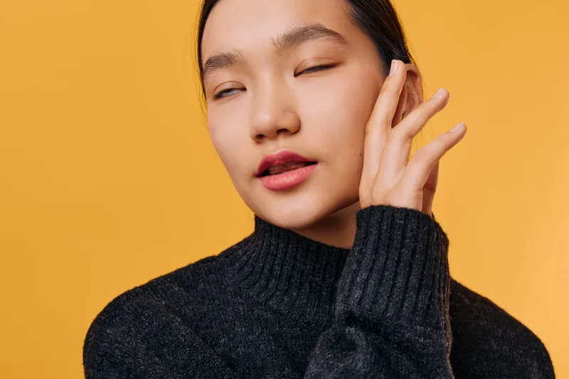 Young woman with closed eyes in black sweater poses against orange background, showing calm expression and hand near face for relaxed and thoughtful mood portrait.
