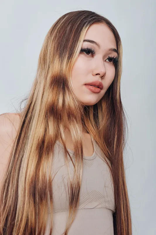 Young woman with long straight hair in beige top looking away with soft expression against plain background. Studio portrait of female model with natural makeup and smooth skin.