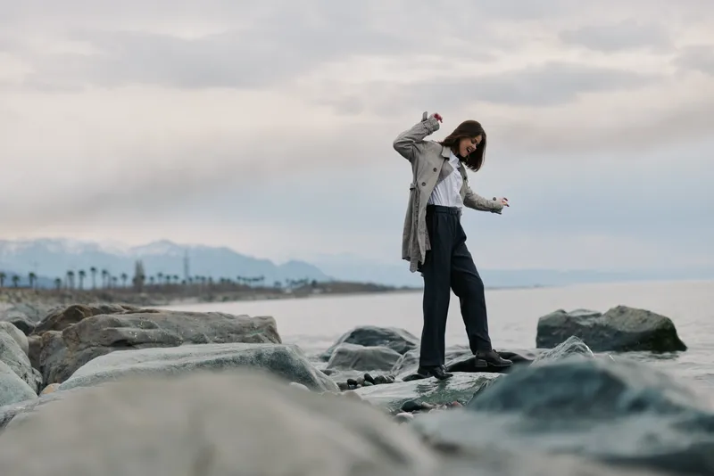 Young woman in casual clothes balancing on rocks by the sea during cloudy weather. Concept of adventure, freedom, and outdoor lifestyle with natural coastal scenery.