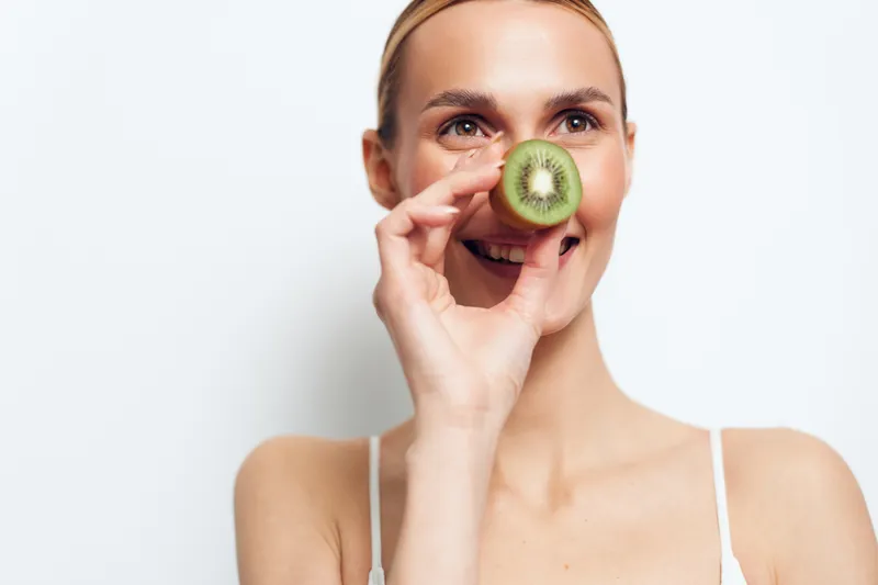 Young woman with blonde hair holds a kiwi slice to her nose in a bright studio portrait, smiling softly against a clean background and natural light, conveying fresh playful energy