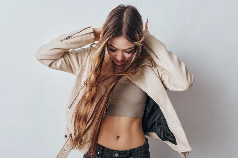Young woman with long hair wearing beige jacket and crop top standing against gray background adjusting her hair with hands above head, casual fashion style.