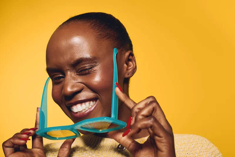 Happy African woman with dark skin smiling joyfully while holding turquoise glasses on yellow background. Portrait of positive young female model with short hair and red nails.