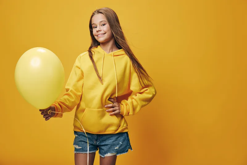 Young girl in yellow hoodie holding balloon, smiling and posing against yellow background. Casual outfit with denim shorts for happy childhood and celebration mood.