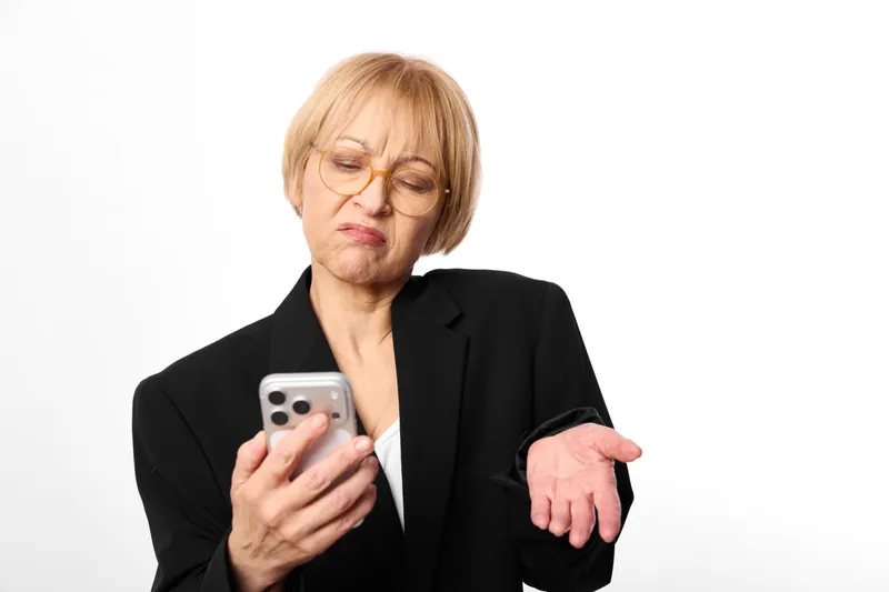 Stressed senior woman with glasses in black business blazer looks worried, holding smartphone, pointing downward with the other hand, isolated on clean white background