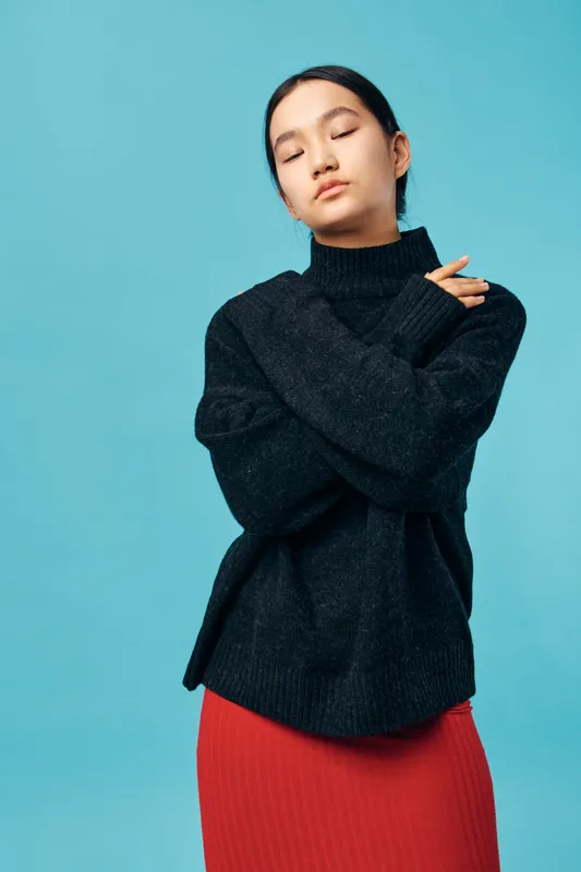 Young woman with closed eyes wearing a black sweater and red skirt standing against blue background. Portrait of calm female with crossed arms and relaxed expression in studio.