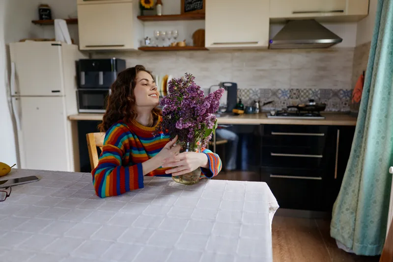 A woman in a striped sweater sits at a kitchen table, admiring a bouquet of lilacs. A warm, cozy home moment reflecting everyday joy, self care, and relaxed lifestyle.