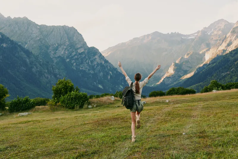 A woman with a backpack stands in a grassy meadow, arms raised in triumph, with expansive mountains behind. Concept: freedom, exploration, and active lifestyle.
