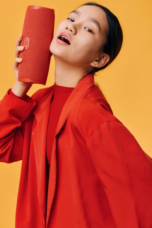 Young woman with red jacket holding red portable speaker near her face singing or enjoying music on yellow background. Fashionable person with casual style and modern technology device.