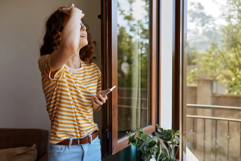 A woman stands indoors by a window, looking outward with a phone in hand. Bright daylight highlights a casual, relaxed home scene, conveying everyday style and self expression.