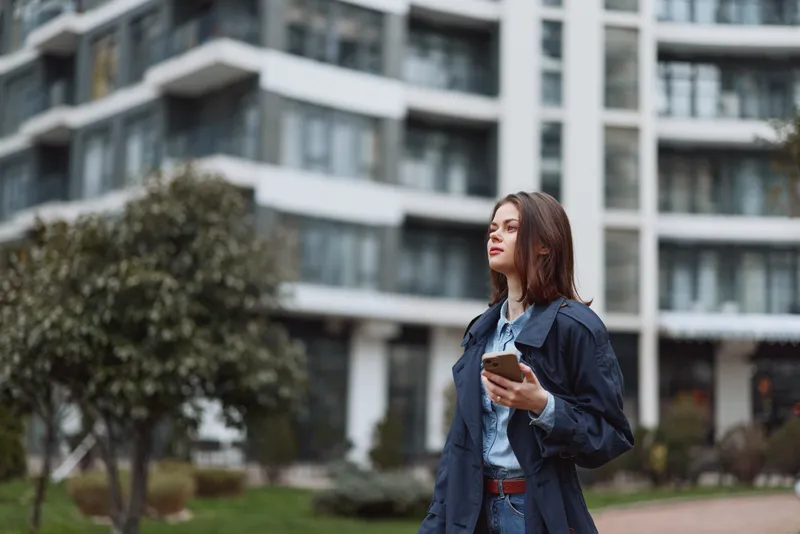 Woman in a denim jacket outdoors, looking up while scrolling on a smartphone. Urban campus setting with modern buildings conveys connectivity, daily routine, and casual style.