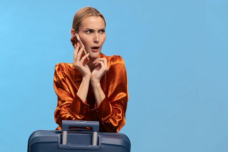 Woman in orange blouse talks on phone with worried expression while standing behind blue suitcase. Concept of travel stress, urgent phone call, and unexpected situation at airport or station.
