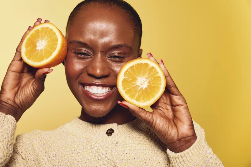 Happy African woman with dark skin holds two orange halves near face, smiling with closed eyes. Yellow background and cozy beige sweater create warm, fresh, and healthy mood concept.