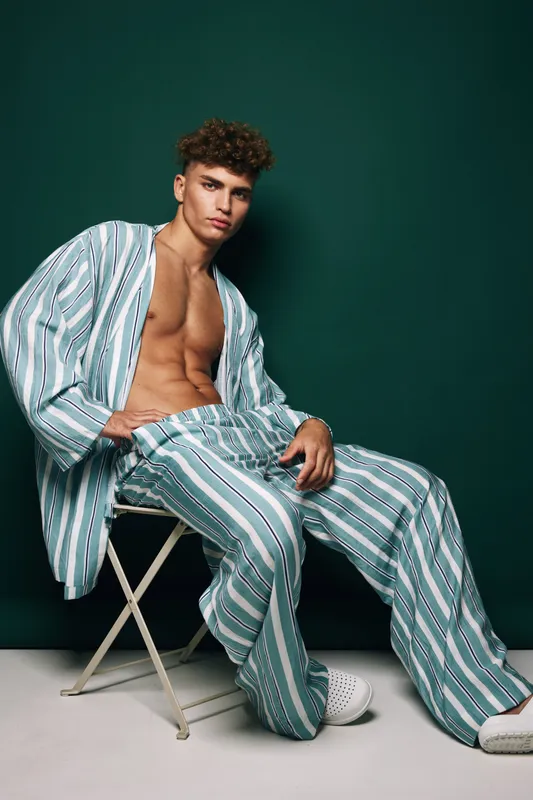 Young man sitting on chair with striped shirt worn open and matching striped pants, exposing his toned chest in studio with dark green background and stylish casual shoes.
