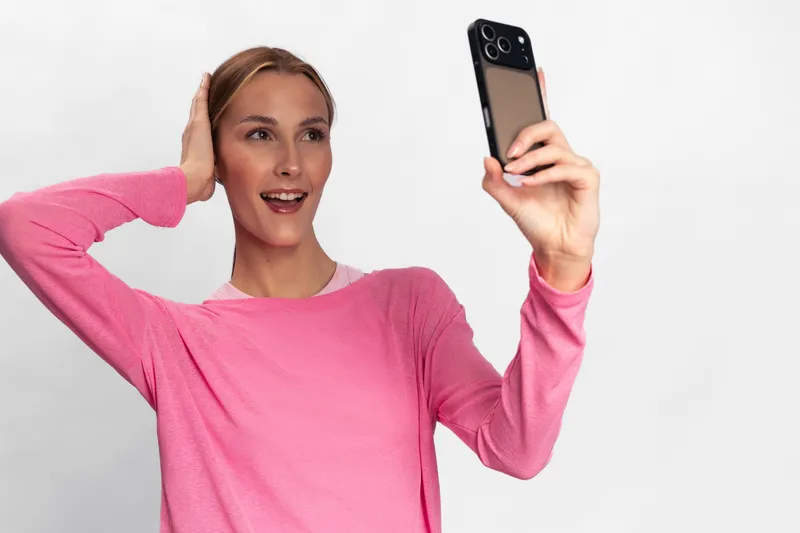 Young woman in pink sweater takes selfie with smartphone on white background. She smiles and touches her hair, expressing joy and casual lifestyle with modern technology concept.