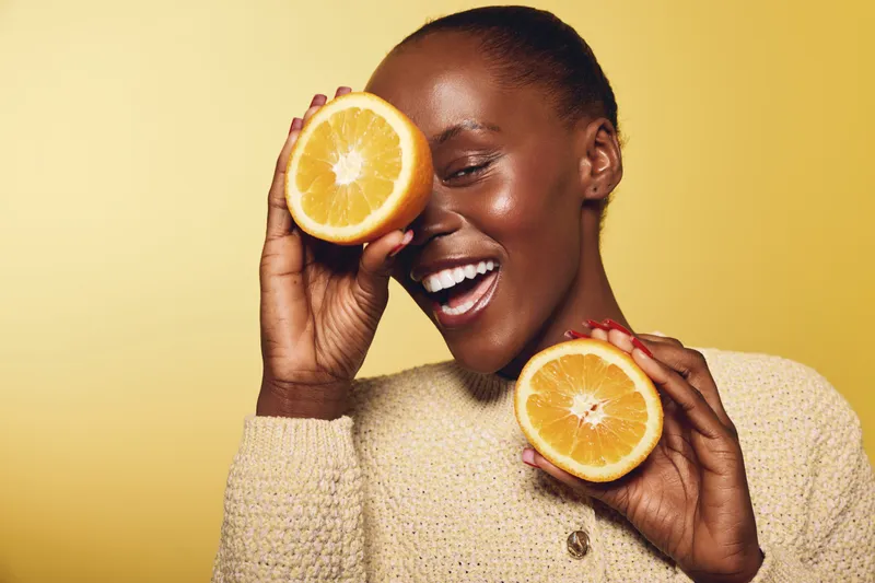 Happy black woman with dark skin holds two orange halves near face smiling joyfully on yellow background. Concept of healthy lifestyle, fruit, vitamin, and natural beauty.