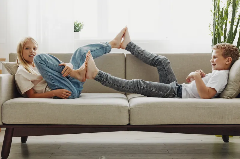Two happy children lying on a beige sofa playfully touching feet in a bright living room with natural light and green plants. Concept of childhood fun, sibling bonding, and home comfort.