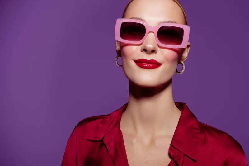 Stylish woman with pink sunglasses and red lipstick poses confidently against purple background. Fashion concept with bold colors and modern accessories.