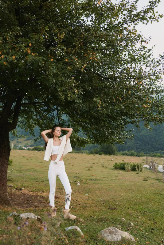 Young woman in white outfit stands under a large tree in a green field with mountains in the background, enjoying a peaceful moment outdoors.
