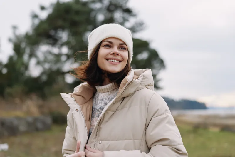 Woman outdoor smiling in a cozy jacket and coat, portrait against a natural scenic background with trees and water, conveying relaxed lifestyle and casual fashion.