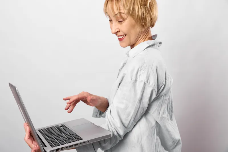 Smiling senior woman using a laptop computer in a studio, shown in side view, wearing light blouse, comfortable posture, looking at screen with cheerful expression today