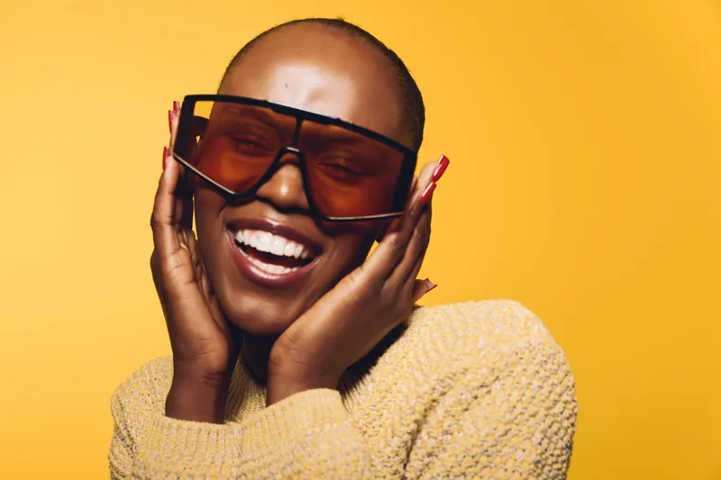Happy black woman with dark skin wearing large sunglasses and a cozy beige sweater smiles joyfully on bright yellow background. Concept of fashion, style, and positive mood.