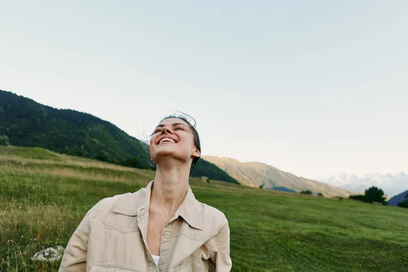 Smiling woman outdoors in a light jacket stands in a grassy field with distant mountains, soaking in sunlight and fresh air. Casual lifestyle, nature, and wellbeing captured in a warm, relaxed mood.