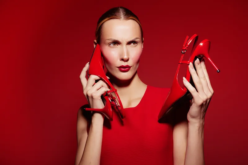 Woman in red dress holds red high heels near face with confused expression on red background. Concept of fashion choice, style dilemma, and footwear selection.