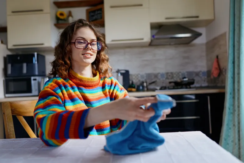 Joyful woman at home folds a blue garment in a bright kitchen, wearing a striped sweater, creating a warm casual lifestyle moment, cheerful and relaxed indoors.